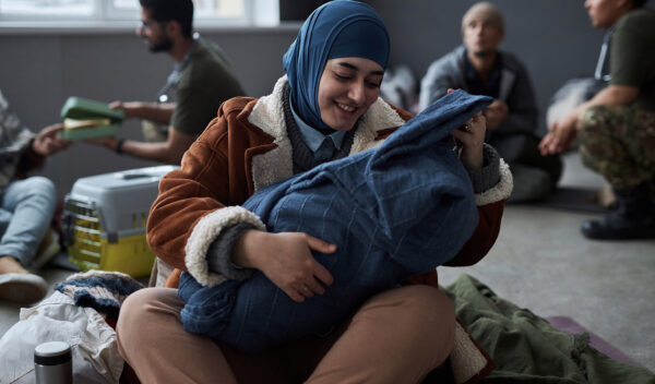 Front view portrait of young Middle Eastern woman playing with baby sitting on floor at refugee shelter, copy space